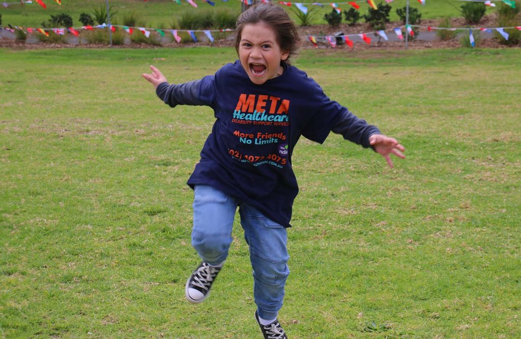 A joyful child running on grass wearing a META Healthcare t-shirt.