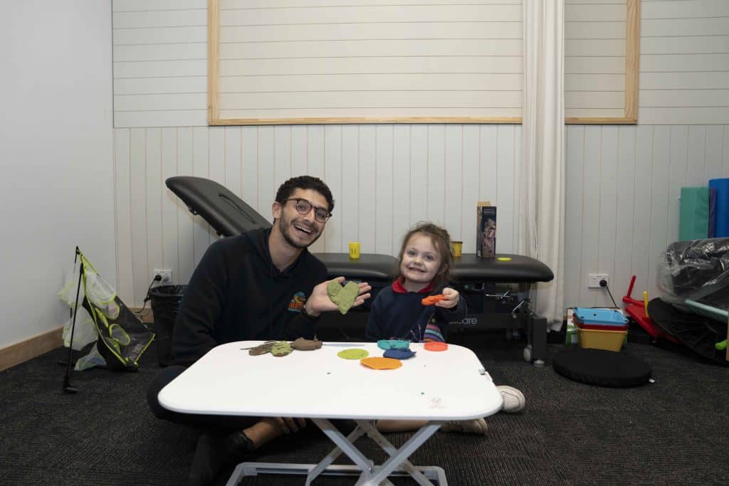 A smiling adult and child playing with colorful modeling clay in a therapy room.