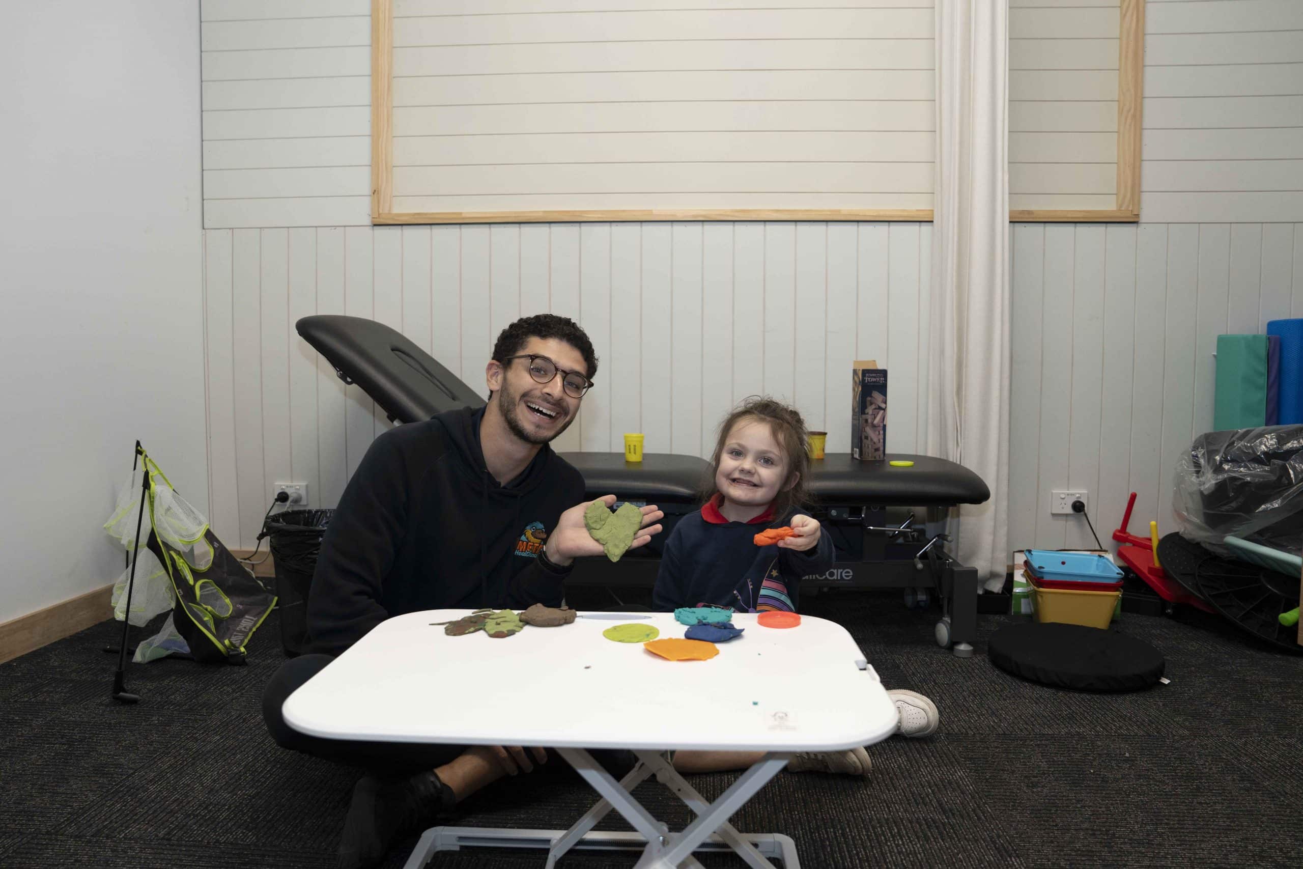 A smiling adult and child playing with colorful modeling clay in a therapy room.