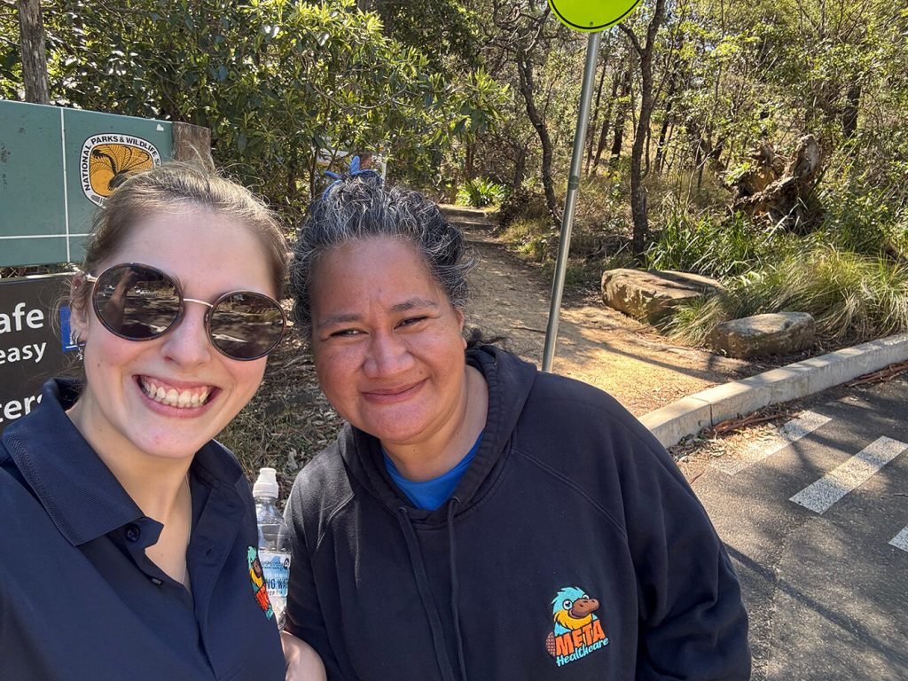Two women smiling in a park setting with a wildlife sign in the background.