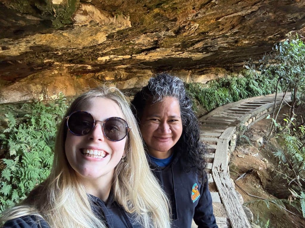 Two friends smiling on a nature trail with lush greenery.
